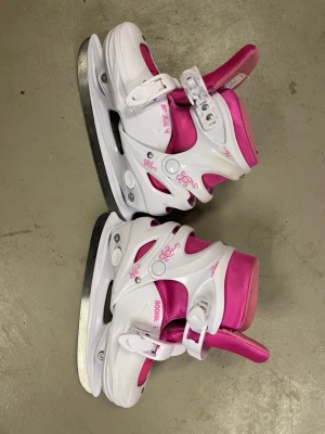 Vita och rosa skridskor Rookie - Snygga vita och rosa skridskor från Rookie med justerbara spännen och vadderad insida för extra komfort. Skridskorna har dekorativa rosa detaljer och är perfekta för isaktiviteter. Passar dig som vill sticka ut på isen.