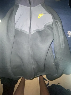 Svart Nike Tech Fleece hoodie - Nike Tech Fleece hoodie i svart och grått med dragkedja framtill och gul logga på bröstet. Har en snygg ficka med dragkedja på ärmen och klassisk huva. Perfekt för dig som gillar sportig och clean stil.