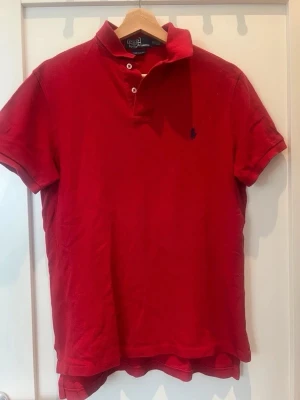 Röd pikétröja från Polo Ralph Lauren - Klassisk röd pikétröja från Polo Ralph Lauren med korta ärmar och krage. Tröjan har knäppning framtill och den ikoniska broderade loggan på bröstet. Använd fåtal gånger. Kom Dm vid frågor!