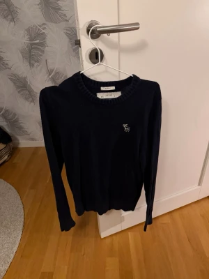 Mörkblå stickad tröja Abercrombie - Mörkblå stickad tröja från Abercrombie & Fitch med rund halsringning och diskret logga broderad på bröstet. Tröjan har långa ärmar och ribbade muddar vid ärmslut och nederkant. Perfekt för lager-på-lager-stil. Nypris 1300