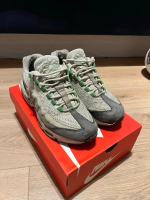 Nike Air Max 95 x Cactus Plant Flea Market - Nike Air Max 95 x Cactus Plant Flea Market sneakers i grått och grönt. Ovandel i mesh och mocka med gröna detaljer på snörningen och en broderad blomma på plösen. Synlig Air Max-sula och unika lager på sidan. Perfekt för dig som gillar streetwear och unika samarbeten.