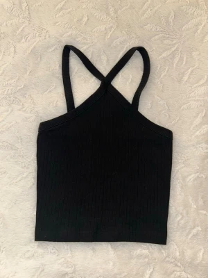 Svart ribbad linnetopp - Snygg svart ribbad linnetopp med smala axelband som korsas i ryggen. Toppen är croppad och har en enkel, stilren design som passar perfekt till jeans eller kjol. Materialet känns mjukt och stretchigt för en bekväm passform.