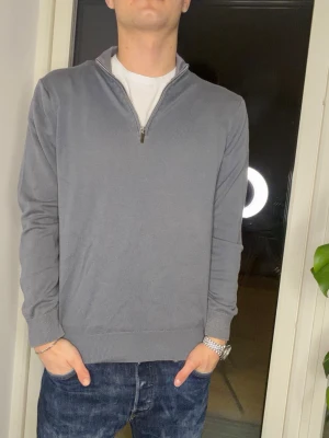 Grå halvzip tröja - En stilren grå tröja med halv dragkedja i kragen. Tröjan har långa ärmar, ribbade muddar och är tillverkad i ett mjukt, tunt material. Perfekt för lager på lager och enkel att matcha med jeans eller chinos.