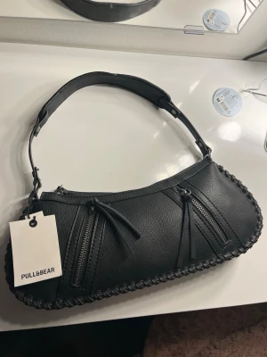 Svart axelväska från Pull&Bear - Trendig svart axelväska från Pull&Bear i konstläder med två dragkedjor framtill och flätad kant. Väskan har ett kort axelband och innerfack med dragkedja. Perfekt till dig som gillar minimalistisk och stilren design.