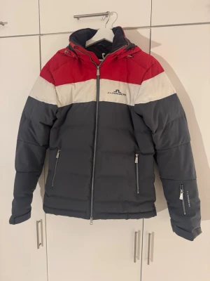 J.Lindeberg pufferjacka röd/grå - Snygg pufferjacka från J.Lindeberg i rött, vitt och mörkgrått. Jackan har huva, dragkedja framtill och två sidofickor med dragkedja. Perfekt för kalla dagar och har en sportig look med färgblock och logga på bröstet.