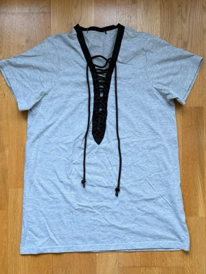 Grå t-shirt med snörning i halsen - Snygg grå t-shirt med svart snörningsdetalj i v-ringad hals. T-shirten har korta ärmar och är tillverkad i mjuk bomull, perfekt för en avslappnad och trendig look. Oversized.