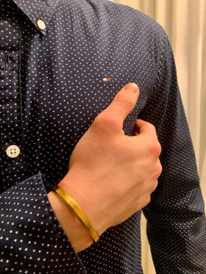 Mörkblå prickig skjorta Tommy Hilfiger - Klassisk snygg mörkblå skjorta från Tommy Hilfiger med vita prickar och klassisk button-down krage. Skjortan har vita knappar och diskret logga på bröstet. stilrena detaljer och tidlös design. Storlek 176cm