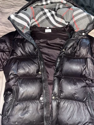 Svart pufferjacka från Burberry - Svart pufferjacka från Burberry med klassiskt rutigt foder i grått, vitt och rött. Jackan har dragkedja framtill, två fickor med dragkedja och hög krage. Perfekt för kalla dagar och ger en stilren look små hål vid sidorna av fickorna annars inget större