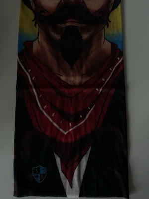 Färgstark halsvärmare med comboy motiv - Cool halsvärmare med tryck av en tecknad mustaschprydd man i rött bandana-mönster. Färgerna går i rött, svart, blått och gult med vita detaljer. Perfekt för dig som vill sticka ut när du åker skidor eller fiskar! 