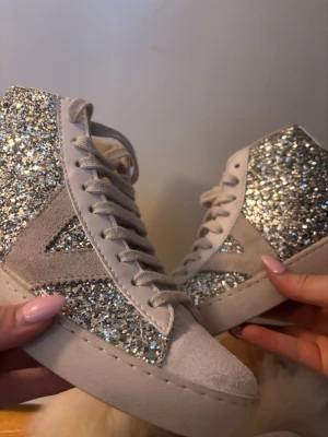 Glittriga höga sneakers i silver - Säljer ett par riktigt snygga sneakers med högt skaft, täckta av silverglitter och detaljer i ljus mocka. Skorna har vit sula och snörning framtill. Perfekta för dig som vill sticka ut och älskar bling!