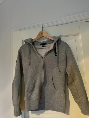 Grå zip hoodie från Ralph Lauren - Snygg grå hoodie från Ralph Lauren med dragkedja, huva med dragsko och fickor framtill. Stort svart logotryck på bröstet och en svart '3' patch på ena ärmen. Perfekt för en avslappnad och sportig stil. Mjuk bomullsblandning för extra komfort.