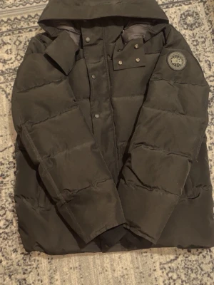 Svart dunjacka från Canada Goose - Säljer en svart dunjacka från Canada Goose med huva och tydlig logotyp på ärmen. Jackan har quiltad design, tryckknappar och dragkedja framtill. Perfekt för kalla dagar och levereras med originalpåse.