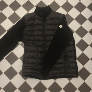 Svart dunjacka från Moncler - Snygg svart dunjacka från Moncler med quiltad front och släta ärmar. Jackan har dragkedja framtill, ståkrage och Moncler-logga på ärmen. Perfekt för kalla dagar och ger en clean, modern look.