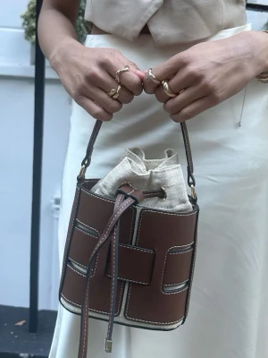Brun bucket handväska med snörning - Snygg bucket handväska i brunt skinn med kontrasterande vita sömmar och beige tyginsats. Väskan har ett coolt rutmönster i lädret och stängs med snörning upptill. Perfekt accessoar för att lyfta din outfit.