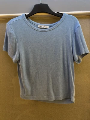Blå croppad t-shirt från Zara - En enkel blå croppad t-shirt från Zara med rund halsringning och korta ärmar. Tillverkad i mjuk bomull som känns skön mot huden. Perfekt till jeans eller kjol för en avslappnad look.