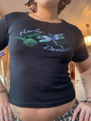 Svart Harley-Davidson t-shirt - Svart t-shirt från Harley-Davidson med tryck av en trollslända och logga på bröstet. Modellen är figurnära med rund hals och korta ärmar. Perfekt för dig som gillar coola vintage-vibbar och snygga tryck.