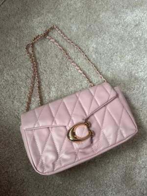 Rosa quiltad axelväska med kedja - Säljer en ljusrosa quiltad axelväska med guldfärgad kedjerem och snyggt spänne framtill. Väskan har ett elegant rutmönster och är tillverkad i syntetmaterial. Perfekt för dig som vill ha en trendig och stilren accessoar.