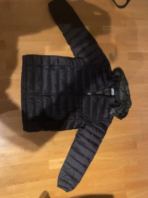 Svart pufferjacka från Stone Island - Säljer en svart pufferjacka från Stone Island med ikonisk patch på ärmen. Jackan har huva, dragkedja framtill och quiltad design. Perfekt för kalla dagar och riktigt snygg streetstil. Passar dig som vill ha en clean och modern look.