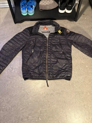 Svart vindjacka från Parajumpers - Snygg svart vindjacka från Parajumpers med gul detalj vid kragen och patch på ärmen. Jackan har dragkedja framtill, två snedställda fickor med dragkedja och quiltad design. Perfekt för dig som vill ha en lätt och stilren jacka.