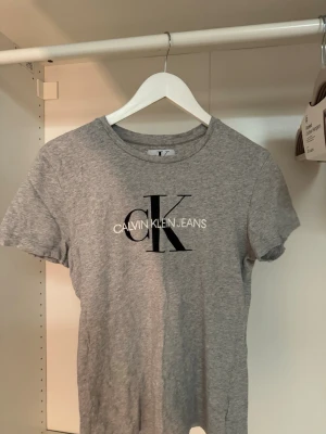 Grå Calvin Klein Jeans t-shirt - Snygg grå t-shirt från Calvin Klein Jeans med klassisk logga i svart och vitt framtill. T-shirten har rund halsringning och korta ärmar. Tillverkad i mjuk bomull för en bekväm känsla och passar perfekt till jeans eller shorts.🤍