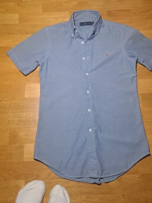 Blå kortärmad skjorta från Ralph Lauren xs - Klassisk blå skjorta från Ralph Lauren med korta ärmar och knappar framtill. Skjortan har button-down krage och den ikoniska broderade loggan på bröstet. Tillverkad i mjuk bomull som känns skön mot huden. Perfekt för en clean och stilren look.