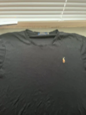 Svart t-shirt från Polo Ralph Lauren - Klassisk svart t-shirt från Polo Ralph Lauren med rund halsringning och den ikoniska broderade polospelaren på bröstet. Tillverkad i mjuk bomull som känns skön mot huden. Perfekt basplagg för en stilren och enkel look.