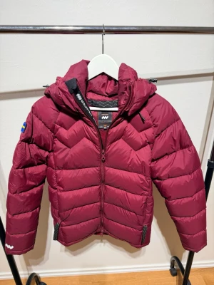 Vinröd dunjacka från Mountain Works - Snygg vinröd dunjacka från Mountain Works med huva och dragkedja framtill. Jackan har quiltad design, två fickor med dragkedja och en svensk flagga på ärmen kommer även med 3 flaggor till och en lagningslapp och knappar. Perfekt för kalla dagar och riktigt skön att bära.