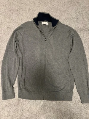 Grå ziptröja från Pier One - Snygg grå ziptröja från Pier One med hel dragkedja och ribbade muddar. Tröjan har en ståkrage med mörkblå insida och två fickor framtill. Perfekt för lager på lager och enkel att matcha med olika outfits.