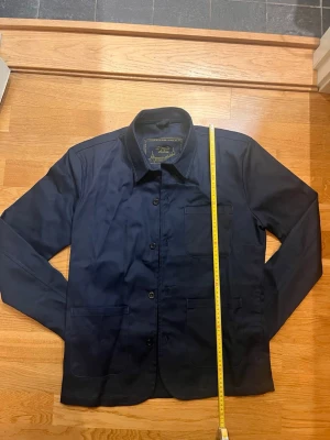 Mörkblå overshirt från donce de la sense  - Snygg mörkblå overshirt från   donce de la sense med klassisk krage, tre framfickor och knäppning framtill. Tillverkad i slitstark bomull som ger en clean och stilren look. Perfekt lager-på-lager-plagg för dig som gillar enkel men genomtänkt stil.