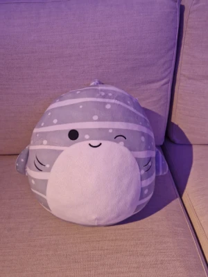 Grå Squishmallow val kudde - Supermjuk och rund kudde från Squishmallows i form av en val. Kudden är grå med vita ränder och prickar, har små fenor och en söt ansiktsdetalj med blinkande öga. Perfekt som mysig inredningsdetalj eller gosedjur. 290 kr ny