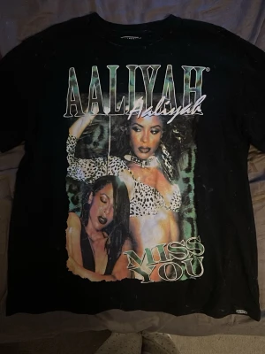 Svart Aaliyah t-shirt med tryck - Trendig  svart t-shirt med stort Aaliyah-tryck på framsidan och texten 'Miss You'. T-shirten har korta ärmar och en avslappnad passform. Perfekt för dig som gillar streetwear och ikoniska motiv. Det svarta tyget är ”uttvättad” i färgen.