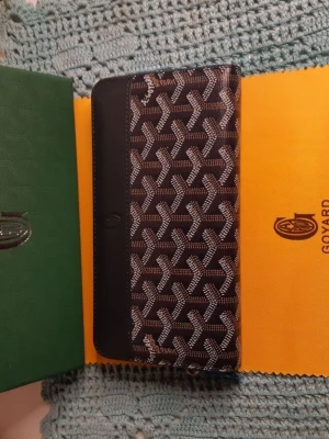 Svart och brun Goyard plånbok - Snygg plånbok från Goyard i svart och brunt med det klassiska Goyard-mönstret. Plånboken har en rektangulär form och är tillverkad i skinn och syntetmaterial. Perfekt för dig som vill ha något både stilrent och ikoniskt.