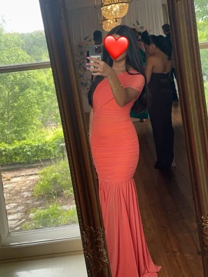 Lång korallfärgad offshoulderklänning - Säljer en lång, figurnära klänning i korall med offshoulder-modell och rynkade detaljer. Klänningen har en volangliknande nederdel och är perfekt för speciella tillfällen. Materialet känns mjukt och följsamt. Det kan va så att de finns fläckar längst nere vid släpet av klänningen. Detta är en XS men passa även nån som bär S eller även M då den är extremt stretchig🥰🩷