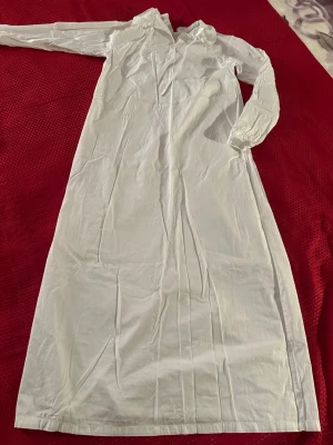 Lucialinne vit storlek 146/152 , längd 120 cm - Klassiskt vitt Lucialinne i lång modell med långa ärmar och spetsdetaljer runt kragen. Knäppning med knapp i nacken och mjukt, tunt tyg som känns skönt mot huden.  Bomull 100%