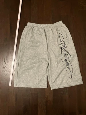 Grå shorts med svart text - Gråa archive shorts med svart tryck längs ena benet. De har resår i midjan och är tillverkade i mjuk bomullsmix. Perfekta för en avslappnad stil och har en loose passform.