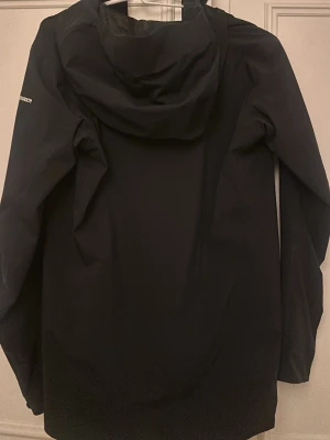 Svart regnjacka från Peak Performance - Svart regnjacka från Peak Performance med huva och hel dragkedja framtill. Jackan har två fickor med dragkedja och är perfekt för regniga dagar. Enkel och stilren design som passar till det mesta.