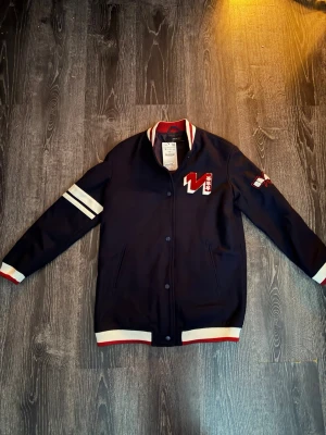 Mörkblå bomberjacka från Zara Basic - Snygg mörkblå bomberjacka från Zara Basic med vita och röda detaljer. Jackan har broderad patch på bröstet och ärmen, ribbade muddar och krage samt knäppning framtill. Perfekt för dig som gillar sportig stil.