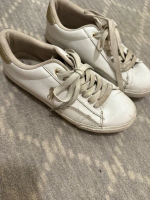 Vita sneakers från Polo Ralph Lauren - Stilrena vita sneakers från Polo Ralph Lauren med gulddetaljer och broderad logga på sidan. Skorna har snörning, rund tå och platt sula. Ovandelen är i skinn och hälen har en glittrig gulddetalj som ger extra touch.