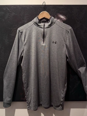 Grå träningströja från Under Armour - Snygg grå långärmad träningströja från Under Armour med halv dragkedja framtill och diskret logga på bröstet. Tillverkad i ett mjukt och stretchigt funktionsmaterial som andas, perfekt för träning eller löpning.