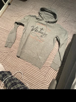 Grå hoodie med snygg text - Snygg grå hoodie från Camp Supply med huva och dragsnören. Framsidan har ett stort vintage-inspirerat tryck i svart och rött med texten 'San Francisco Vintage'. Tillverkad i mjukt bomullsmaterial och har ribbade muddar vid ärmar och nederkant.