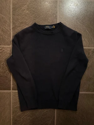 Mörkblå stickad tröja Polo Ralph Lauren - Klassisk mörkblå stickad tröja från Polo Ralph Lauren. Tröjan har rund halsringning, långa ärmar och ribbade muddar vid ärmslut och nederkant. Diskret logga broderad på bröstet. Perfekt till jeans eller kjol för en clean look.