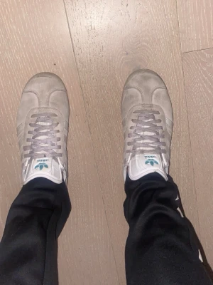 Adidas Samba grå sneakers - Snygga Adidas Samba sneakers i grå mocka med klassiska vita detaljer och snörning. Skorna har den ikoniska Adidas-loggan på plösen och guldfärgad 'SAMBA'-text på sidan. Perfekta för dig som gillar retrostil och streetwear.