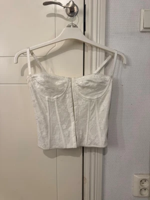 Vit spetskorsett med snörning - Trendig vit korsett med tunna axelband och vacker spetsdetalj. Framsidan har markerade sömmar och bägge kupor är lätt vadderade. Baksidan har snygg snörning som ger en justerbar passform. Perfekt för att lyfta vilken outfit som helst.