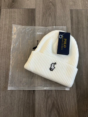 Vit ribbstickad mössa Polo Ralph Lauren - Vit ribbstickad mössa från Polo Ralph Lauren med klassisk uppvikt kant och broderad logotyp framtill. Perfekt accessoar för kyliga dagar och enkel att matcha med olika outfits.
