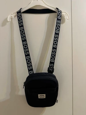 Svart axelväska från Hugo Boss - Snygg svart axelväska från Hugo Boss med justerbart axelband där det står BOSS i vitt. Väskan har en fyrkantig form, dragkedja upptill och en praktisk ytterficka framtill. Perfekt storlek för det viktigaste och stilren design med tydlig logga. Äkta!