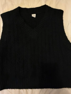 Svart stickad väst med V-ringning - Snygg svart stickad väst med V-ringning och ribbade kanter. Västen har ett klassiskt kabelstickat mönster och är ärmlös, perfekt att bära över en skjorta eller t-shirt. Materialet ser mjukt och lite tjockare ut, vilket ger en mysig känsla.
