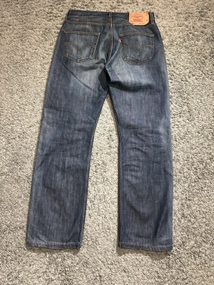 Blå raka jeans från Levi's 501 med unik tvätt - Klassiska blå Levi's 501 jeans med rak passform och femficksmodell. Jeansen har en mörk tvätt och är tillverkade i slitstarkt denimtyg av bomull. Perfekta för en avslappnad och tidlös stil. Storleken på jeansen är 34/34 men passar mer som 32 eller 33 i midja 