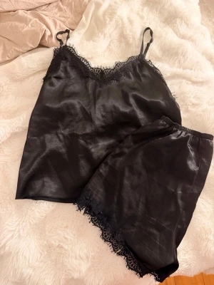 Svart satinpyjamas med spetsdetaljer - Snyggt svart pyjamasset i satin med linne och shorts. Linnet har tunna axelband och spetskant vid ringningen, medan shortsen har elastisk midja och matchande spets vid bensluten. Perfekt för en chill kväll hemma eller mysiga nätter.