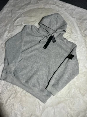 Grå hoodie från Stone Island - Snygg grå hoodie från Stone Island med klassisk huva och dragsko. Hoodien har en stor magficka och den ikoniska Stone Island-loggan på ärmen. Tillverkad i mjukt bomullsmaterial som känns riktigt skönt mot huden. Perfekt för dig som gillar streetwear och enkel stil.