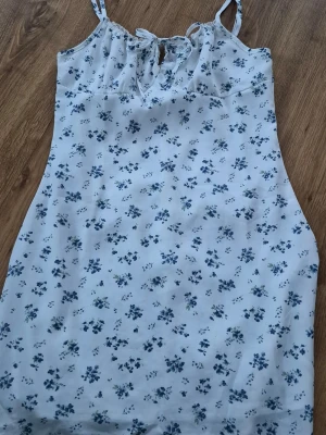 Blommig kortklänning med snörning från shein - Söt kortklänning med blå blommönster på vit botten. Klänningen har smala axelband, snörning framtill och en lätt figurnära passform. Perfekt för varma dagar och sommarens alla tillfällen.